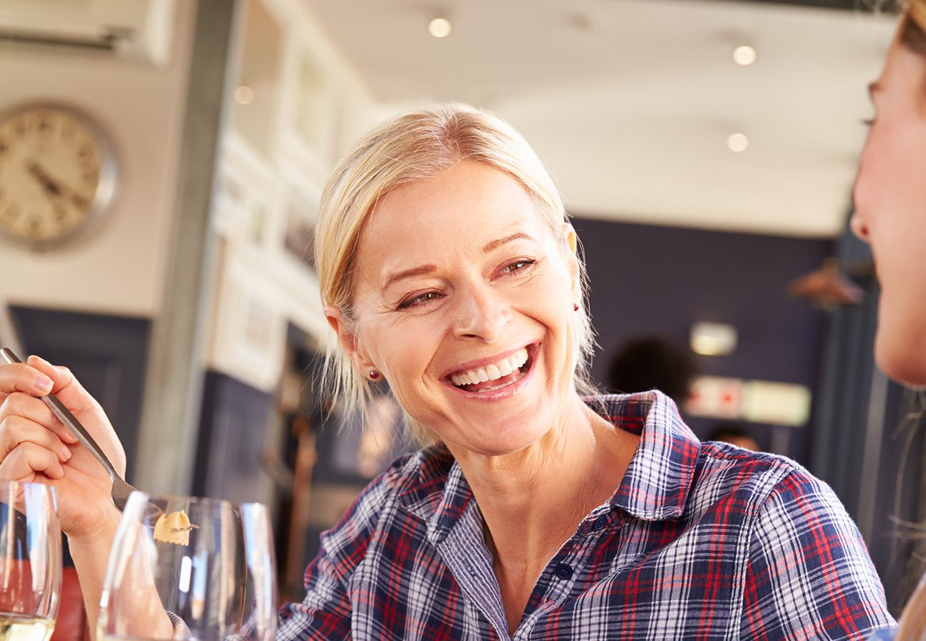 A smiling older woman with blonde hair sits at a table in a restaurant, holding a fork and talking to someone. She wears a plaid shirt and appears happy and engaged. A clock is visible on the wall in the background.