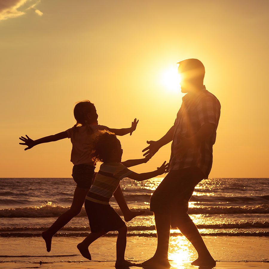 Silhouettes of a man and two children playing joyfully on the beach at sunset, with the sun low in the sky and waves in the background.