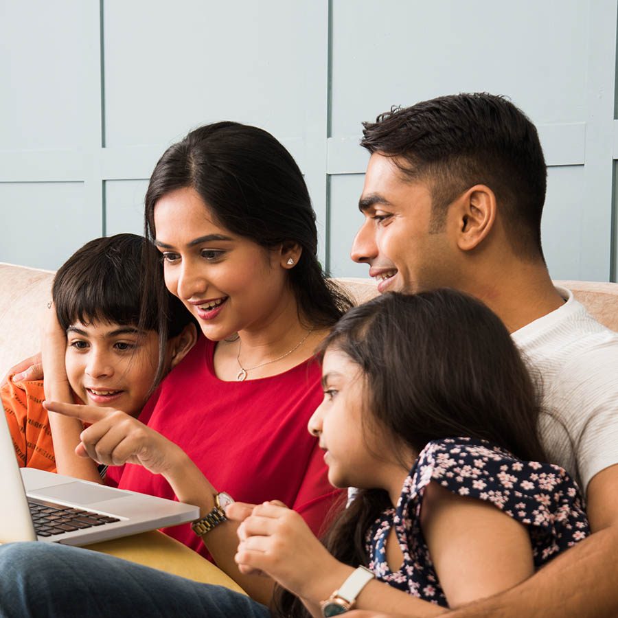 A happy family of four, including a mother, father, son, and daughter, sits together on a couch, smiling and looking at a laptop screen. The mother is pointing at the screen while the children watch attentively.
