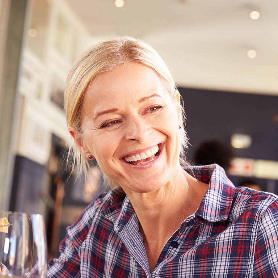 A smiling woman with blonde hair in a ponytail wears a plaid shirt and holds a glass, sitting indoors in a warmly lit setting.