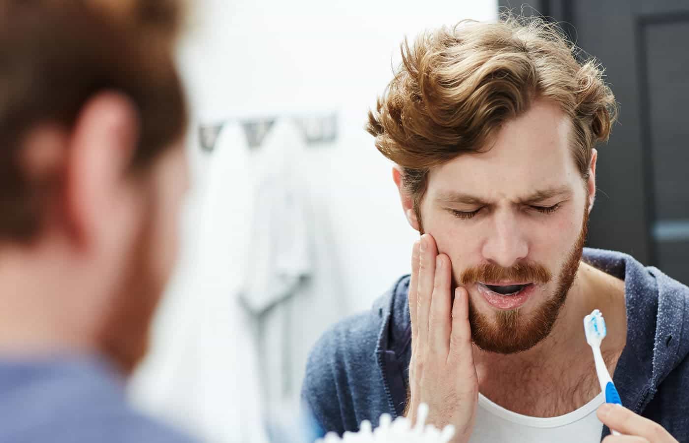 A man with a beard is touching his cheek and wincing in pain, possibly experiencing a toothache or jaw discomfort.