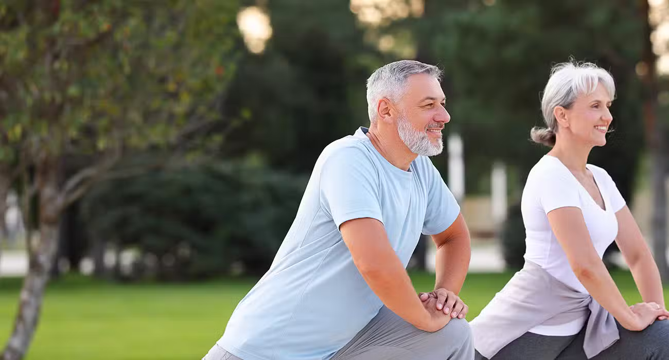 Two older adults, a man and a woman, are exercising outdoors in a park, smiling as they perform lunges on the grass. They appear healthy and active, wearing comfortable athletic clothing.