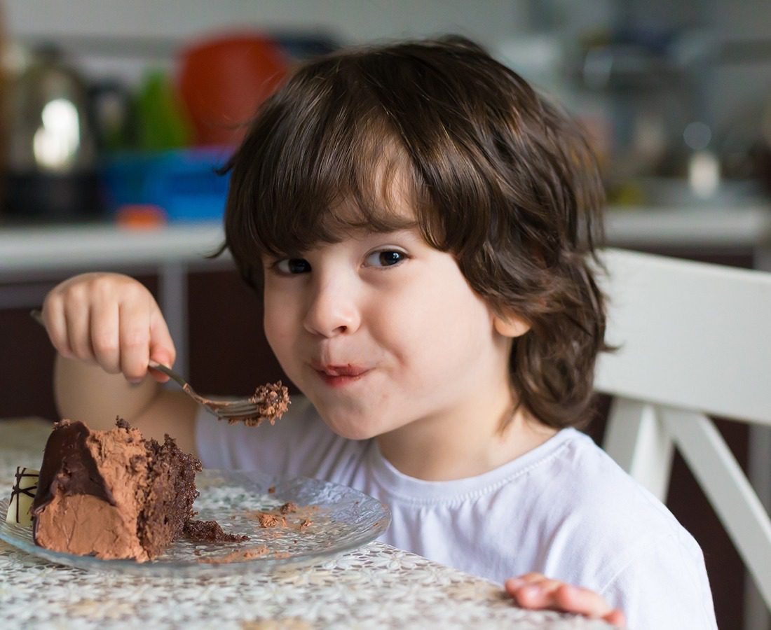 A young child with brown hair sits at a table, smiling while eating a piece of chocolate cake with a fork. The child wears a white shirt and appears happy in a cozy indoor setting.
