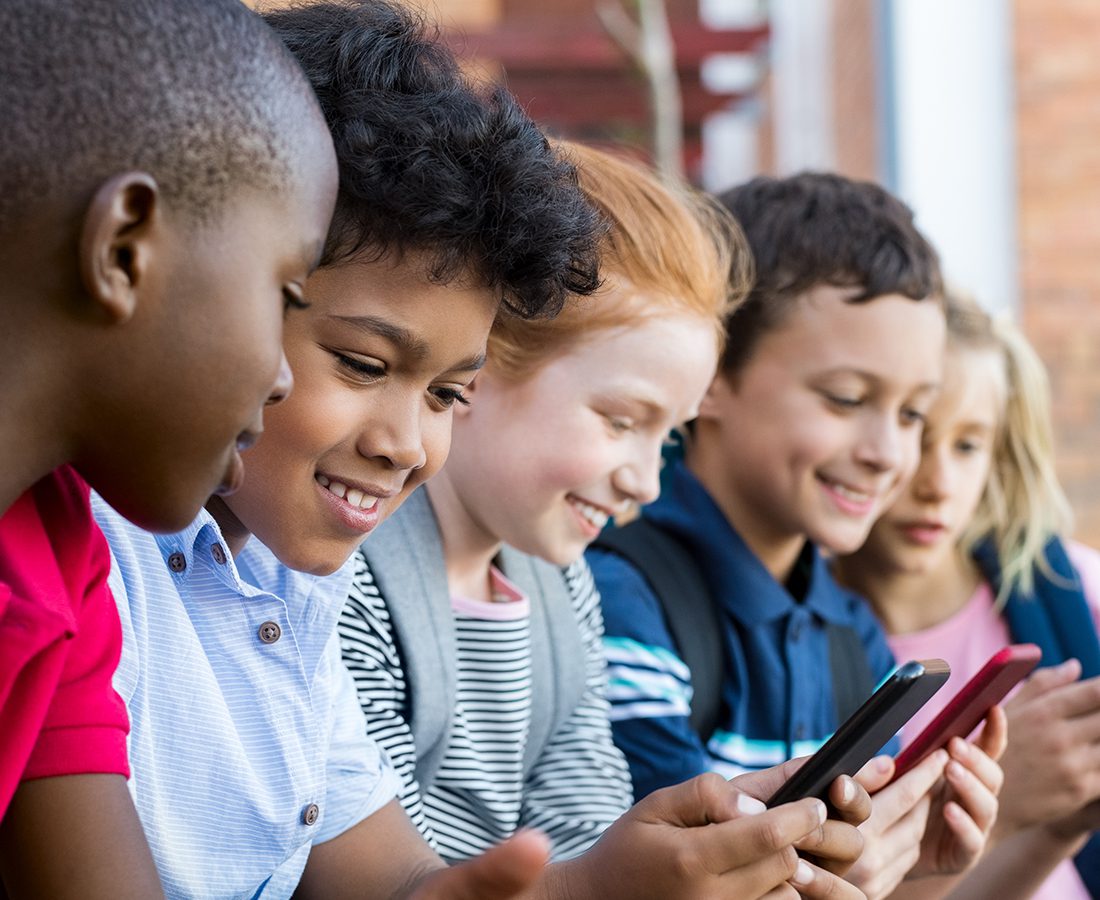 Five children sit closely together outdoors, smiling and looking at smartphones they are holding. They appear to be enjoying themselves and are focused on their screens.